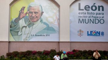 Varias personas se sientan delante de una pancarta colocada sobre una pared con una imagen del Papa Benedicto XVI que dice en español "León está listo para recibir al Papa y el mundo".