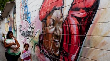 Una mujer y sus hijos camina frente a un mural pintado del presidente Hugo Chávez donde se lee un mensaje en español que dice "Pa' lante Comandante".