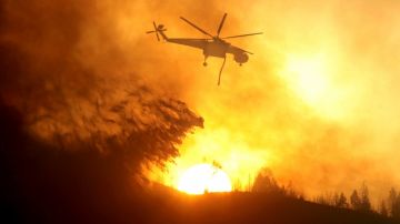 Un helicóptero echa agua sobre las llamas que parecen no dar tregua a los bomberos.  Más personas se han visto  obligadas a dejar sus hogares a concecuencia de la expansión del fuego.