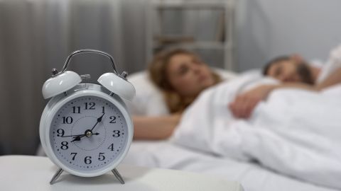 Young,Couple,Sleeping,In,Bed,With,Ringing,Alarm,Clock,On