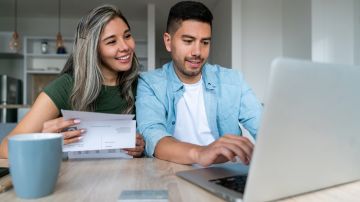 Happy Latin American couple at home paying bills online on their laptop and smiling - financial technology concepts