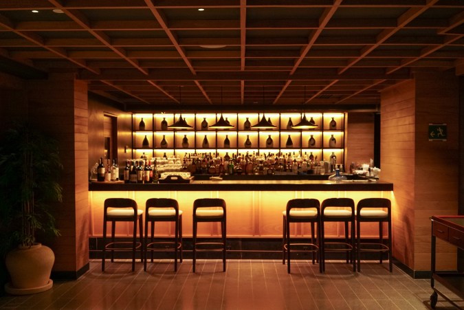 Interior del bar de KOBO en UNICO 20°105°, con estanterías iluminadas, botellas alineadas y sillas altas frente a una barra de madera.