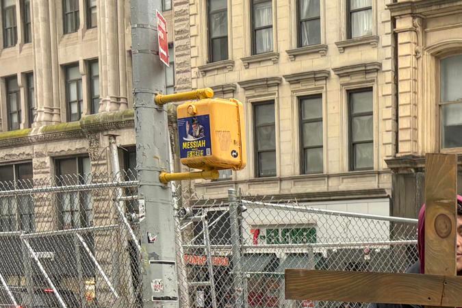 A person holding a wooden cross stands on a city sidewalk, while another person dressed in religious attire looks on. The background includes urban buildings and a partially snowy street.