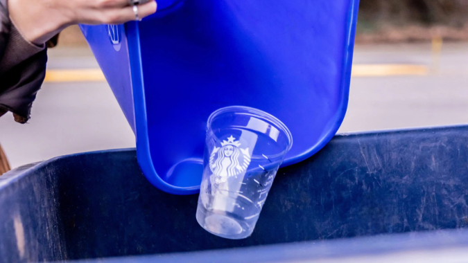 Persona reciclando un vaso de plástico transparente de Starbucks en un contenedor azul, promoviendo hábitos de reciclaje y cuidado ambiental.