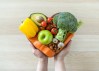 Manos sosteniendo un bowl en forma de corazón lleno de vegetales y frutas sobre fondo gris