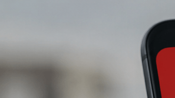 Mano sostiene un celular con una alerta sísmica en pantalla y edificios desenfocados al fondo.
