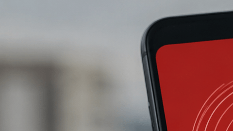 Mano sostiene un celular con una alerta sísmica en pantalla y edificios desenfocados al fondo.