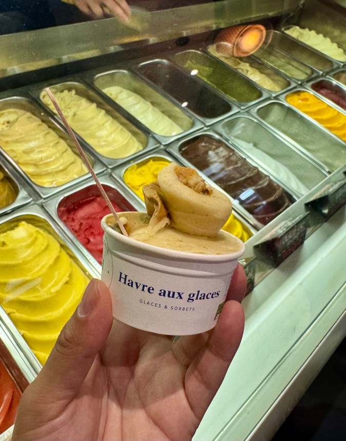 Ficha para WordPress: Alt Text: Vasito de Havre aux Glaces con un helado de tono acaramelado frente a la vitrina de sabores en el mercado Jean-Talon, Montreal