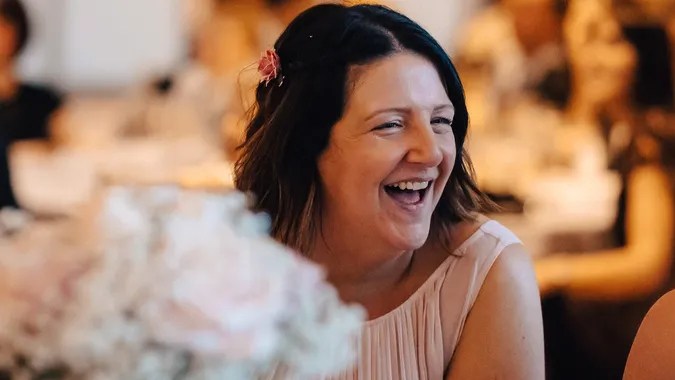 A woman laughing joyfully at a social gathering, wearing a light pink dress. She has dark hair adorned with a small flower and is sitting at a table decorated with flowers in the foreground.