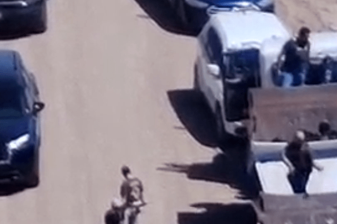 An aerial view of several vehicles, including pickup trucks and police cars, on a dirt road with individuals present near the vehicles. Some people appear to be emerging from the back of the trucks.