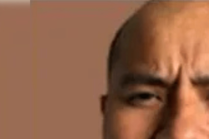 Close-up portrait of a man with a serious expression, featuring a closely shaved head and a short beard, against a plain background.