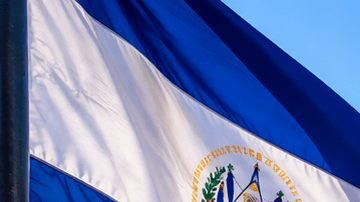 Mujer contempla un lago y un volcán en El Salvador junto a la bandera salvadoreña, en una escena de viaje y regreso al país.